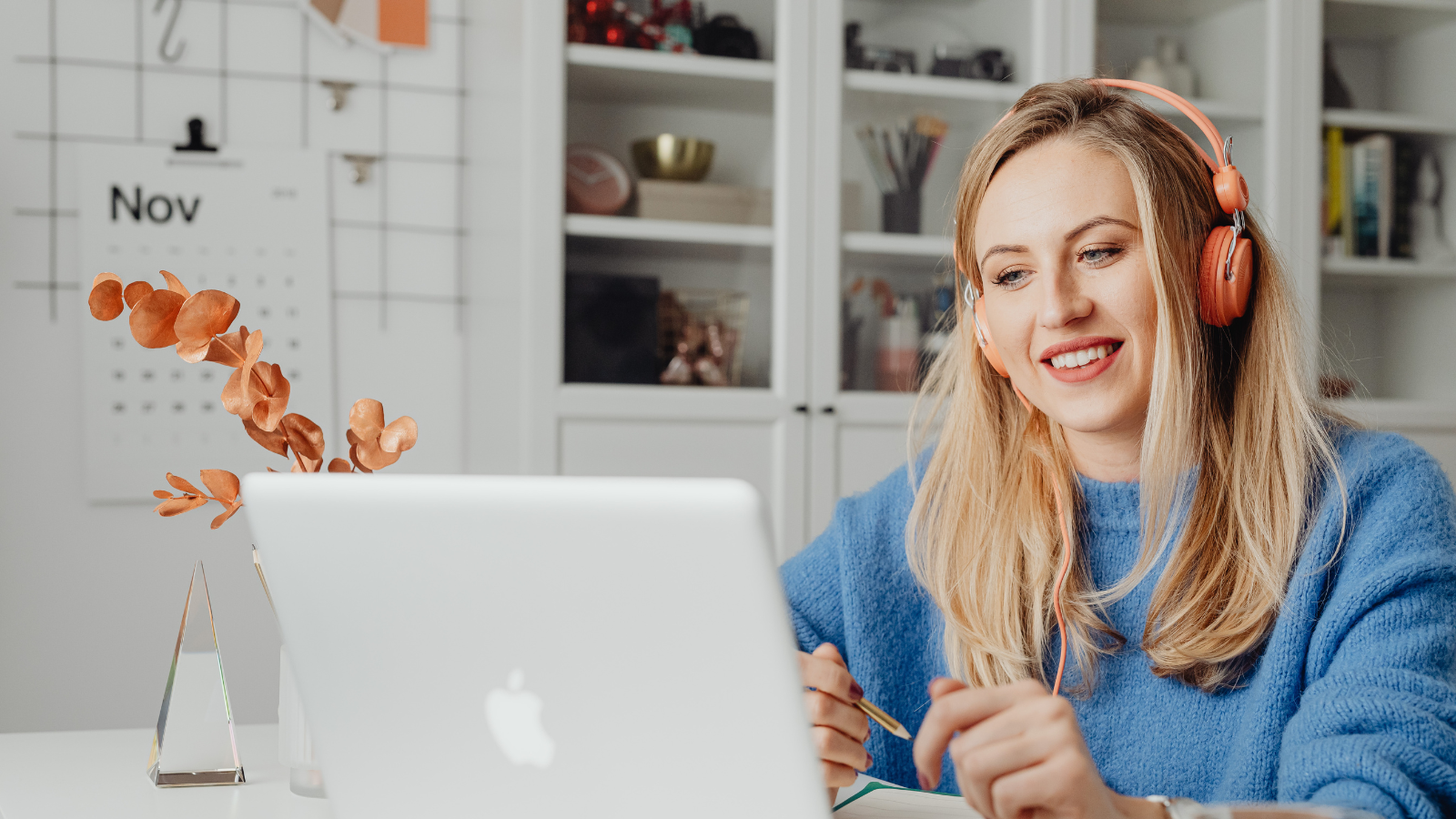 A young woman with long blonde hair, wearing orange headphones and a blue jumper, working at a laptop